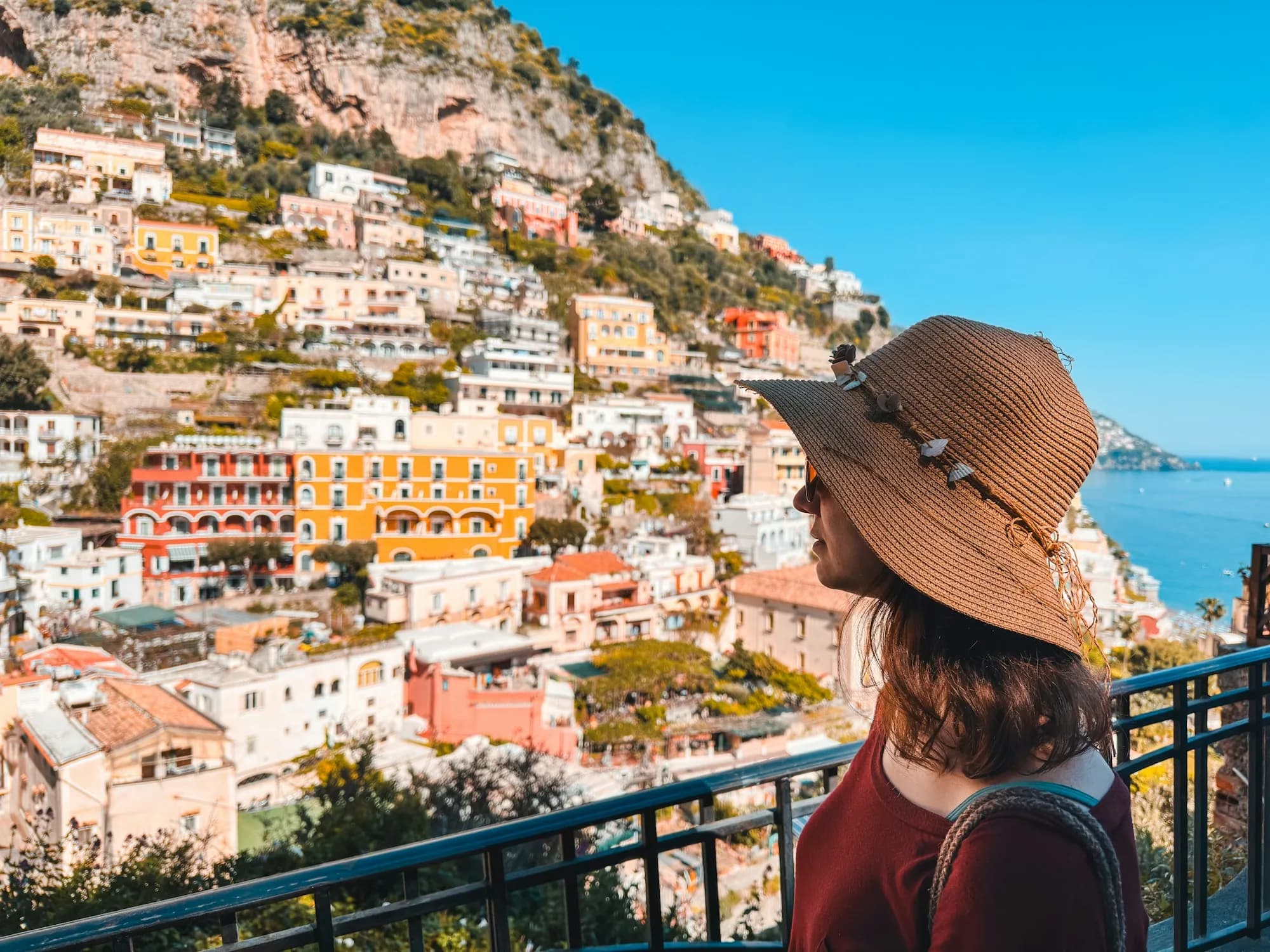Mulher de chapéu de palha observando um vilarejo colorido na encosta e o mar ao fundo